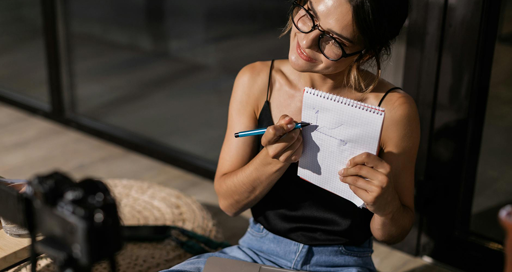 A Woman Sits At A Table With A Notebook And Pen, Drawing A Rough Data Stats Diagram