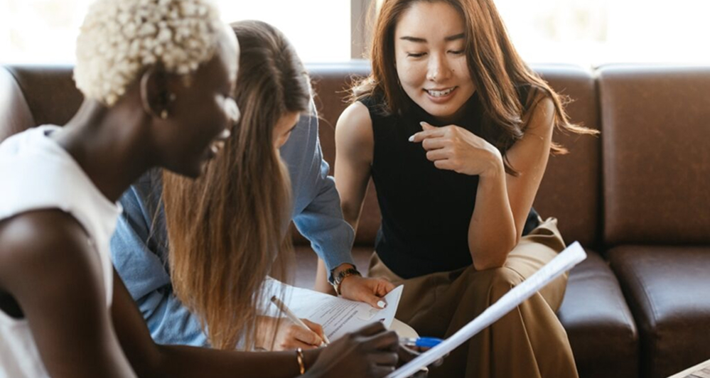 Three Women Sitting On A Couch, Reviewing Documents And Discussing Their Contents.