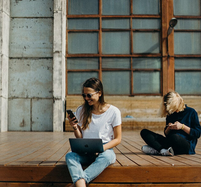 Two Women Sitting On A Wooden Deck, Each Engaged With Their Smartphones